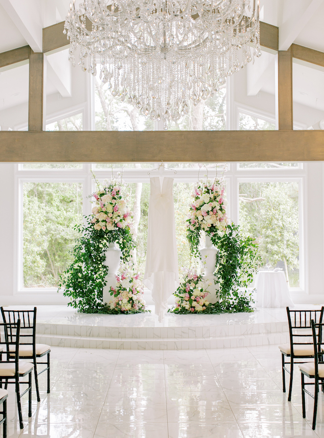 A wedding dress hanging in front of the altar with greenery and pink and ivory florals in the southern-inspired wedding venue.
