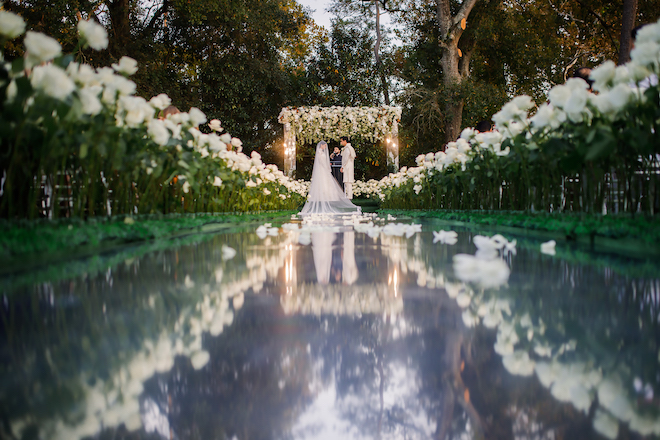 A bride and groom standing at an altar lined with white florals and a mirrored aisle. 