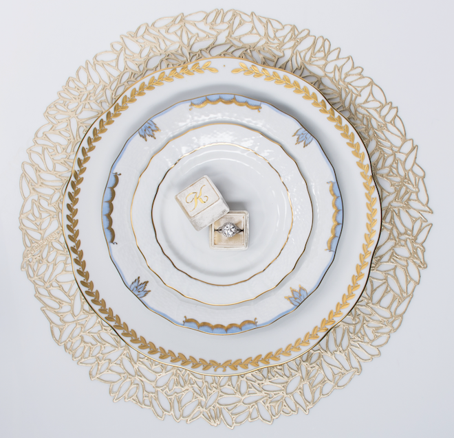 A tabletop set of a gold placemat, a white dinner plate with a gold edge, a white salad plate with blue and gold design and a white bread and butter plate with a gold rim. There is a "K" monogrammed ring box with a diamond ring sitting on the bread and butter plate.