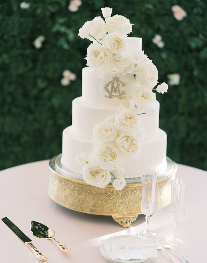 A four-tier white wedding cake with white roses garnishing the front and a gold monogram on the third tier. 
