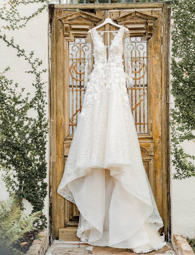 A wedding gown with floral appliques hanging on a brown door with greenery on the walls. 