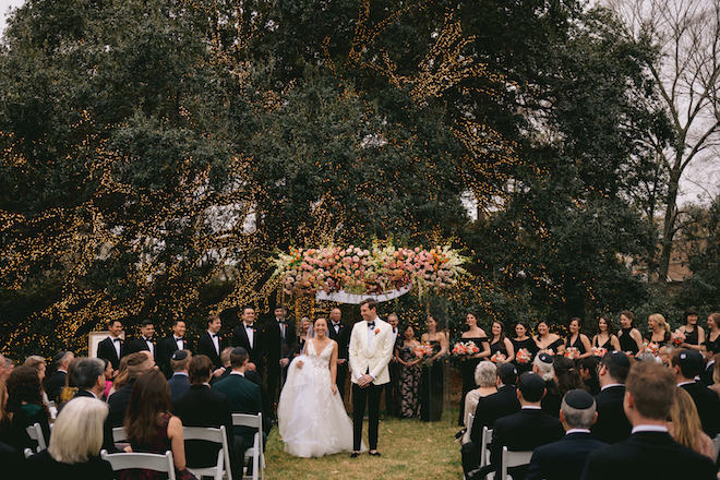 The bride holding her dress and smiling while the groom looks at her. They wedding party is wearing black standing on either side of the floral-filled chuppah with a large lit-up tree behind them.
