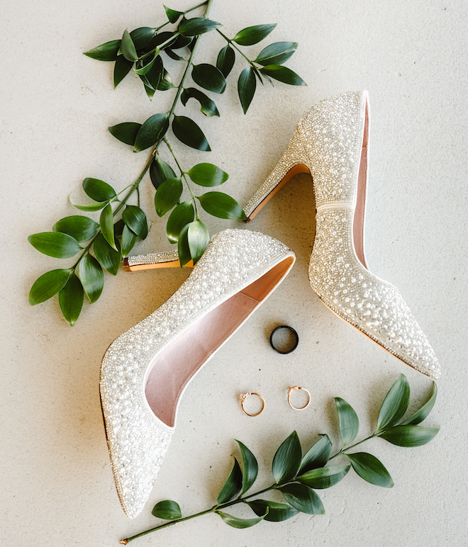 Sparkly white heels next to a black wedding band and gold hoops, with greenery surrounding it. 