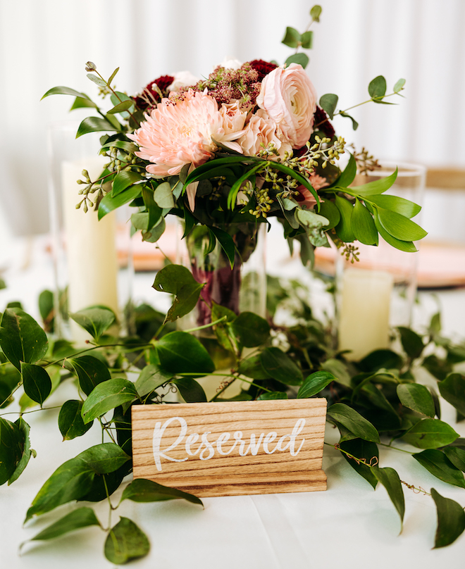 A reception table centerpiece with blush and red florals and greenery with a wooden sign that says "Reserved" in white cursive. 