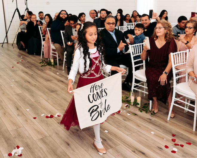 A young girl in a maroon dress walking down the aisle with a sign reading "Here Comes The Bride"