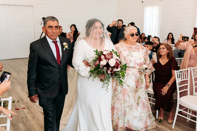 The bride wearing a veil over her head and holding a red and white bouquet with greenery walking down the aisle with her parents on either side of her. 