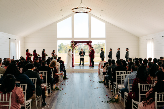 The guests watching the ceremony in a white barn chapel with a red altar. The bride, groom and the wedding party are standing at the altar. 