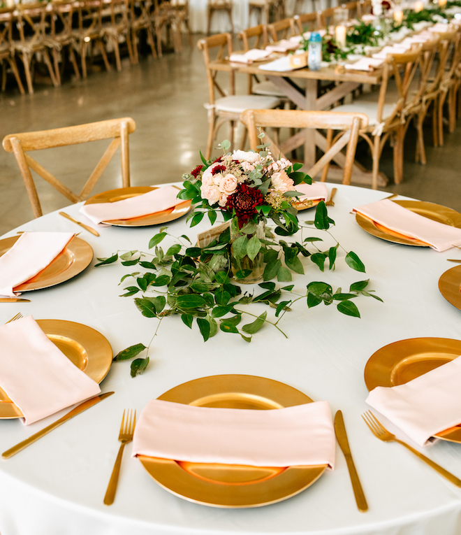 A round table with a greenery centerpiece with blush and red florals. Their are wooden chairs around the round table with gold plates and silverware with blush napkins on the plates. 