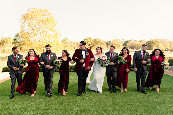 The bride and groom linking arms with their wedding party walking in a grass field. The wedding party is wearing gray suits with red ties and deep red dresses. 