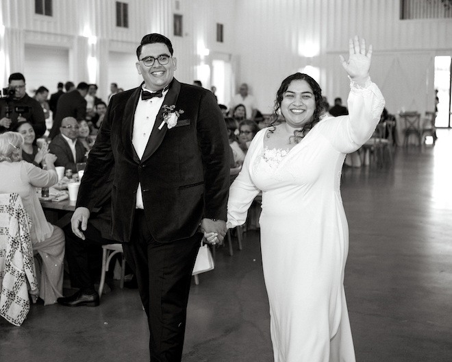 The bride waving to guests and holding hands with the groom walking into their wedding reception. 