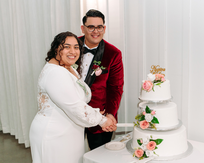 The bride and groom smiling while they cut their three tier white wedding cake with pink and white flowers. 