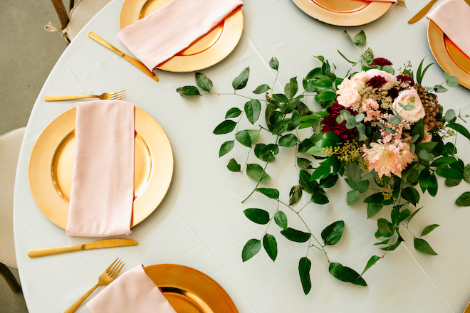 A round table with gold plates and silverware with blush napkins. A centerpiece with greenery, blush and red florals is in the center. 