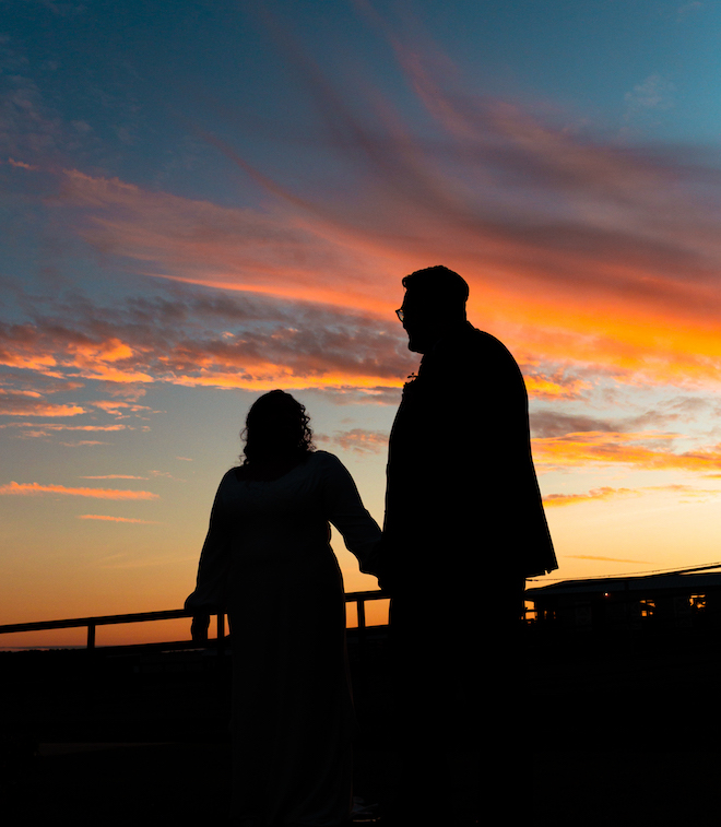 The bride and groom holding hands with an orange and blue sunset in the background.