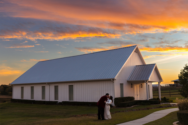 A bride and groom kissing in front of a barn-style wedding venue during sunset.
