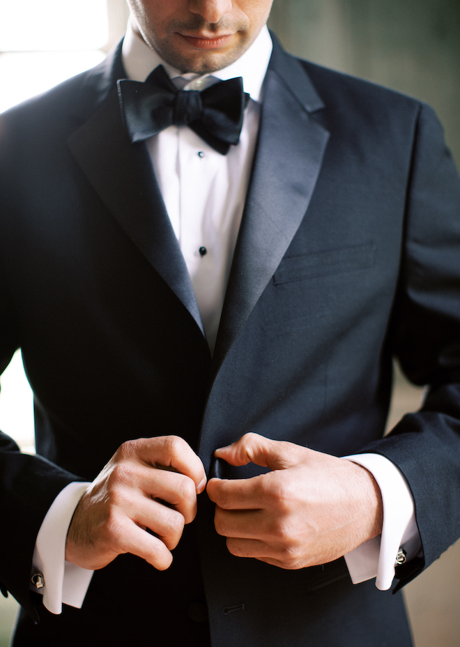 A man wearing a black suit and bowtie buttoning up his suit jacket. 