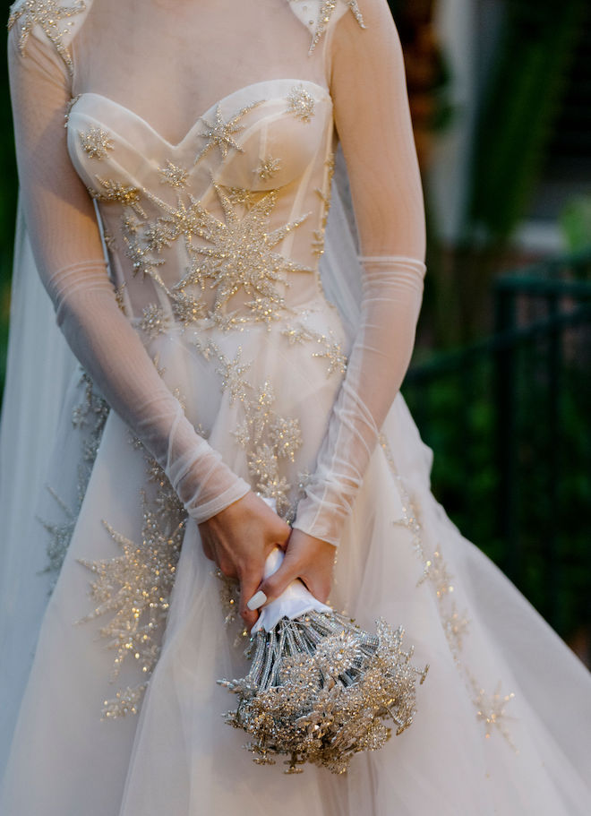 A wedding gown with a corset top and sheer sleeves. The gown has crystal embellishments in the shapes of snowflakes. The bride is holding an all-crystal bouquet. 
