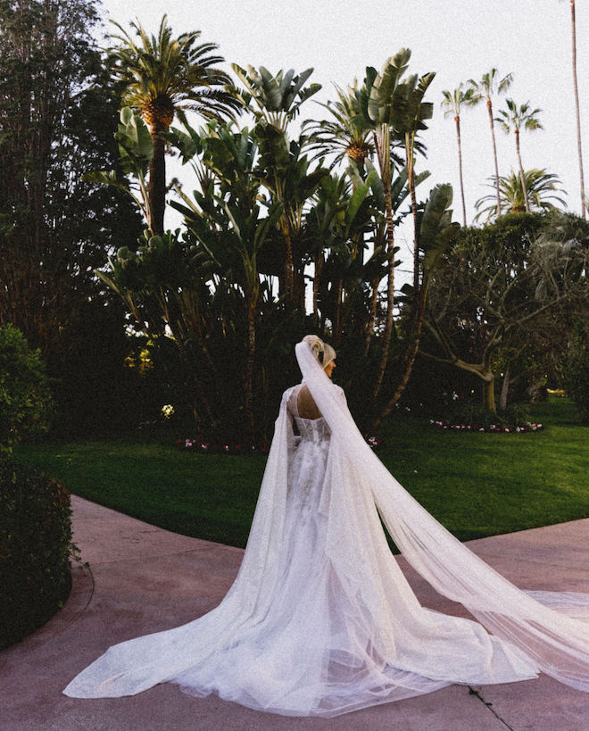The bride facing palm trees and a green lawn, showing off the back of her dress and her veil trailing behind her. 