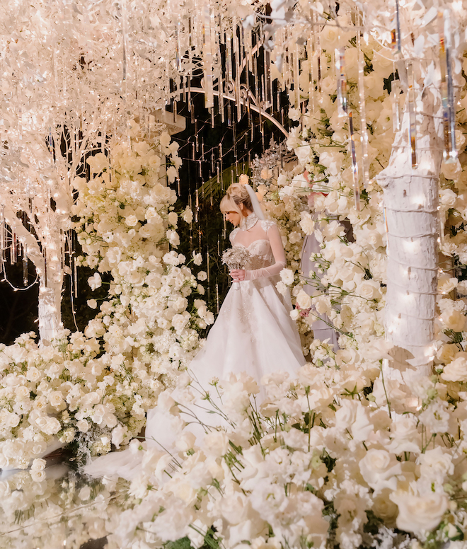 The bride standing under an altar of white flowers and lit-up icicle trees, holding her crystal bouquet. 