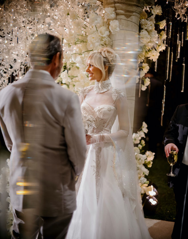 The groom facing the bride as she smiles standing at the altar during their wedding ceremony with white florals and twinkling lights behind her.