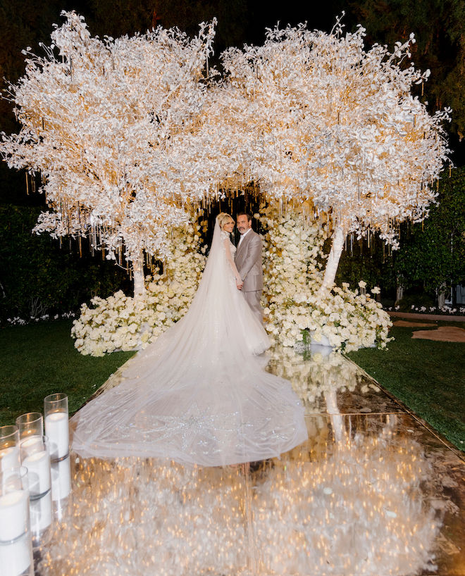 The bride and groom smiling in front of the altar decorated with white florals and lit-up icicle-inspired trees.