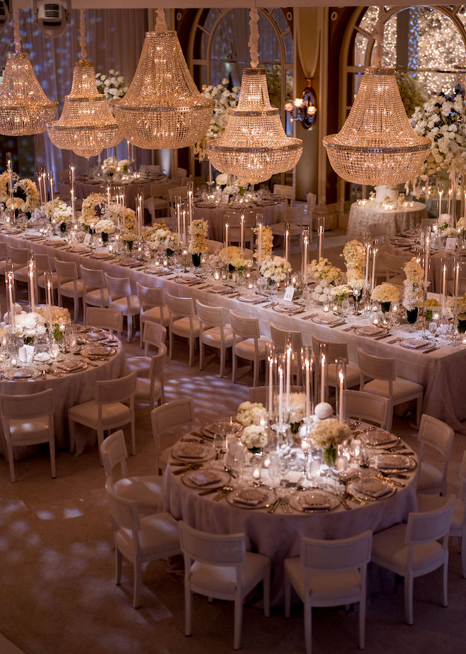 The reception decorated with a long table surrounded by round tables. The tables are decorated with white floral centerpieces and pillar candles. Five large chandeliers hang above the reception tables.