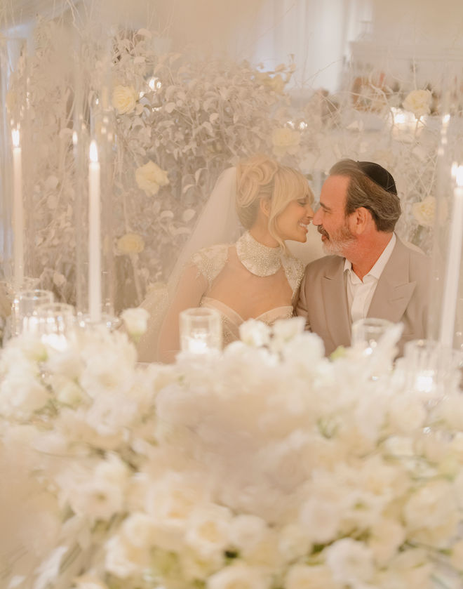The bride and groom sitting at a reception table with white florals and candlelight. The couple are smiling as they are about to kiss. 
