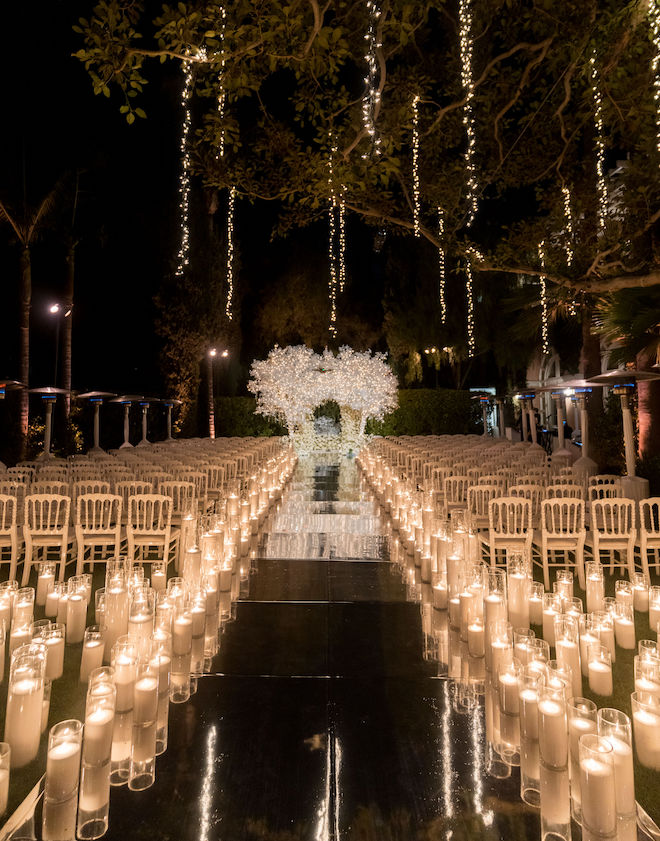 Gold chairs with candles on either side of the mirrored aisle leading to the aisle with icicle-inspired trees and lights.