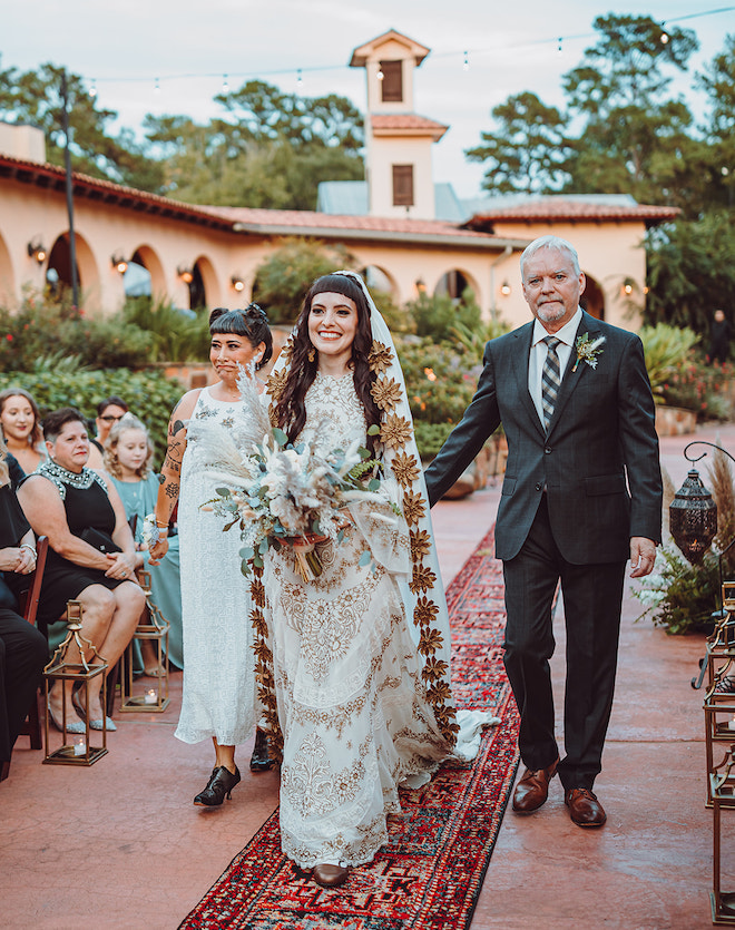 The bride smiling walking down the aisle with her parents walking behind her. She is wearing a Cathedral-style bohemian veil with an ivory and gold detailed gown.