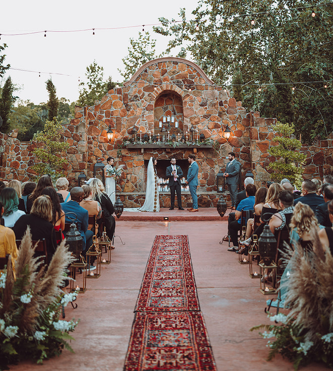 The bride and groom holding hands at the altar at Madera Estates decorated with bohemian-style floral arrangements and a red rug going down the aisle. 