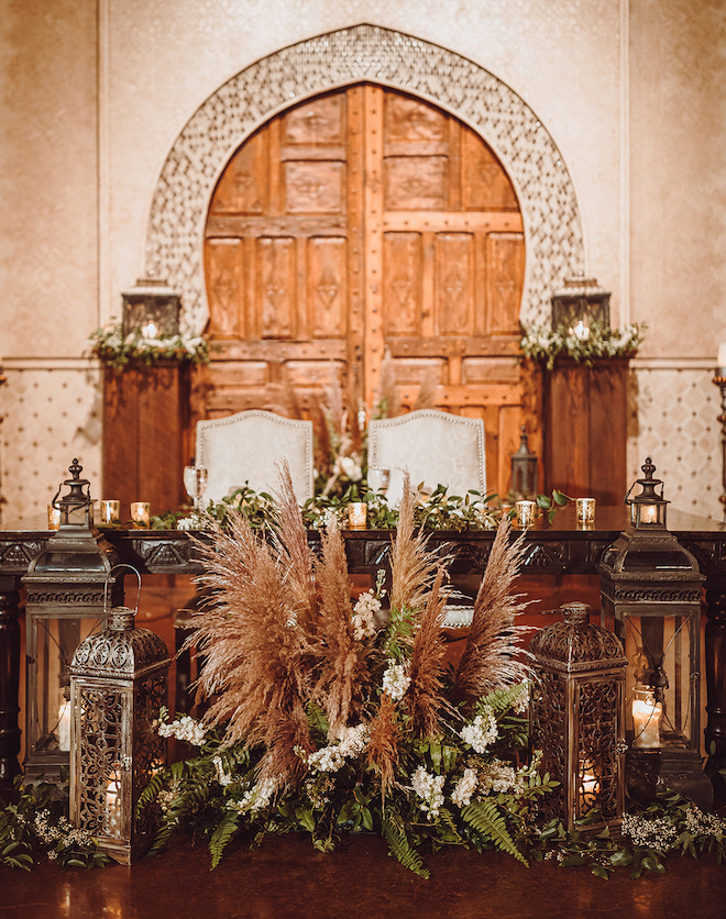 The reception table decorated with pampas grass and greenery with two gray chairs in the middle. 