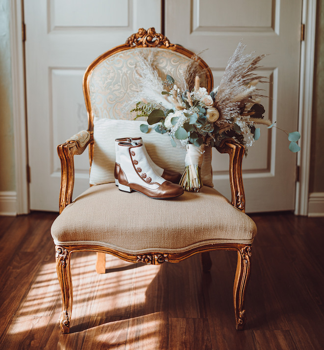 White and brown boots and a bouquet with pampas grass, greenery and white florals in a cream chair. 