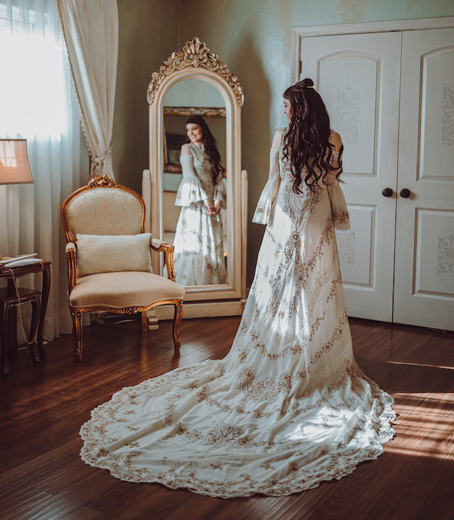 A bride wearing an ivory and gold gown standing in front of a mirror. 