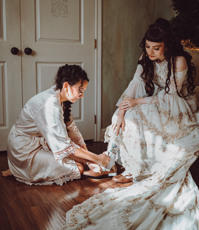 A bridesmaid helping the bride put on her boots wearing her intricate ivory and gold gown. 