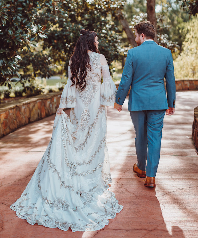 The back of the bride and groom holding hands and the bride pulling up the bottom of her bohemian style ivory and gold gown. 