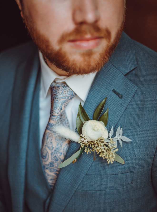 The grooms blue suit with a pink and blue print tie and a white flower with greenery pinned to his suit jacket. 