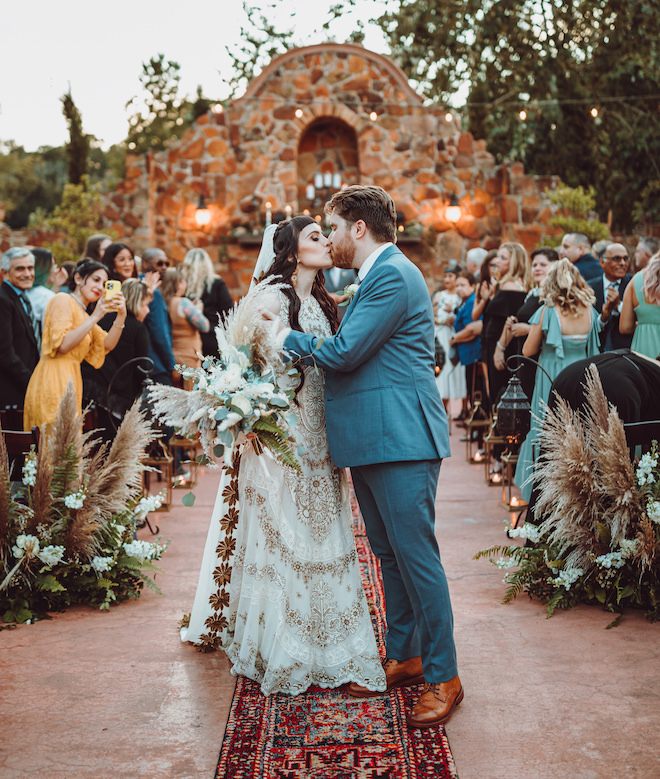 The bride and groom kissing at the end of the bohemian red rug while guests cheer in the background. 