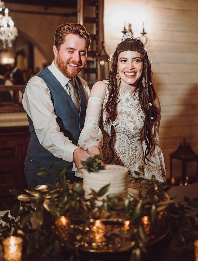 The groom and the boho bride smiling with flowers in her hair as they cut the white cake. 