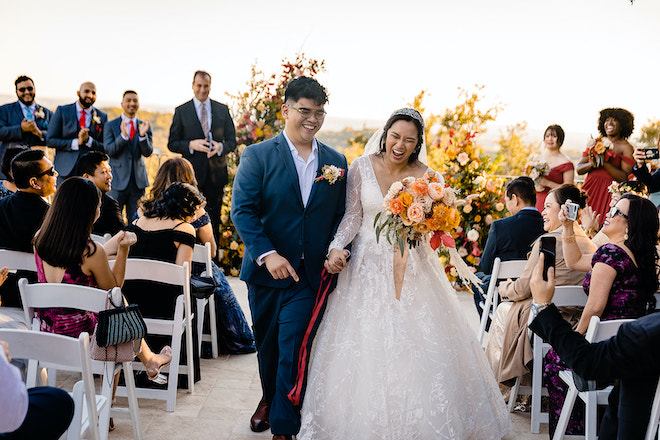 The bride and groom holding hands and laughing as they walk back down the aisle from their alfresco autumn ceremony. 