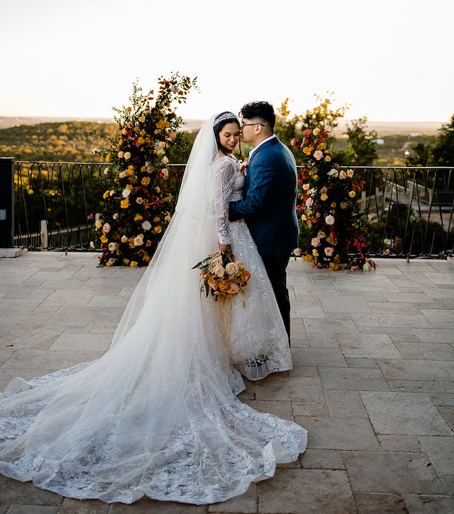 The groom kissing the brides cheek in front of a floral arrangement of greenery and autumn colored flowers with a view of the Austin hill country. 