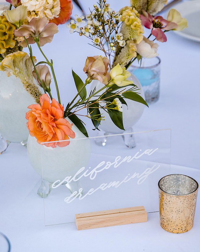 A clear sign with white cursive reading "California dreaming" sitting on a table with a white tablecloth and florals.