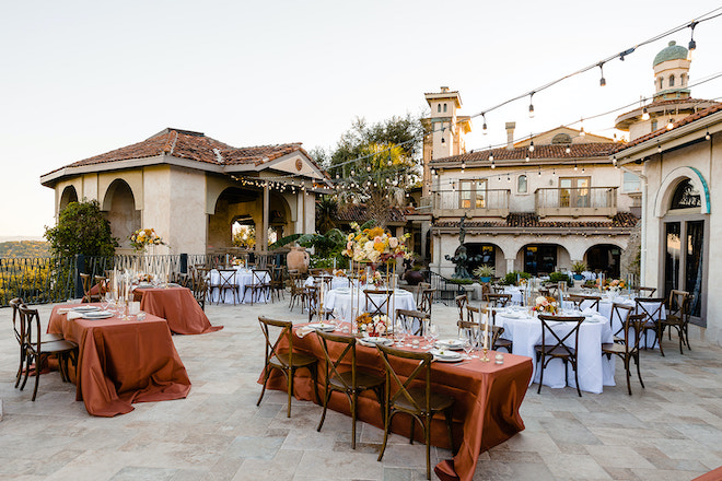 A hill country venue with twinkling lights and orange and white tables with floral arrangements. 