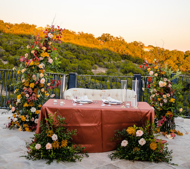A cream loveseat with an orange tablecloth and candles framed by a floral arrangement of greenery and autumn colored flowers with a view of the Austin hill country. 