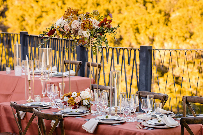 A reception table decorated with a burnt orange tablecloth, clear glassware and floral centerpieces with gold, red and blush flowers overlooking the Austin hill country. 