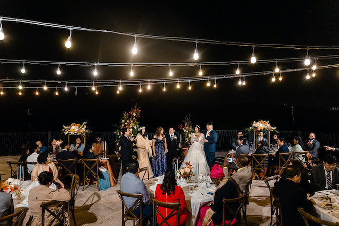 The bride and groom standing with guests during the toasts under the twinkling lights at their alfresco reception. 
