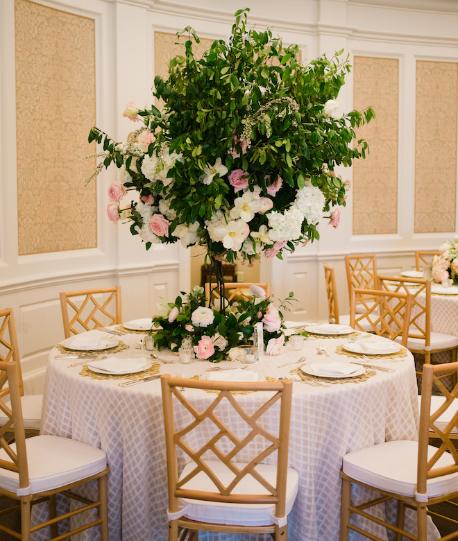 A circular table with a gray and white checked tablecloth with brown chairs and a large centerpiece of greenery and pink and white florals. 