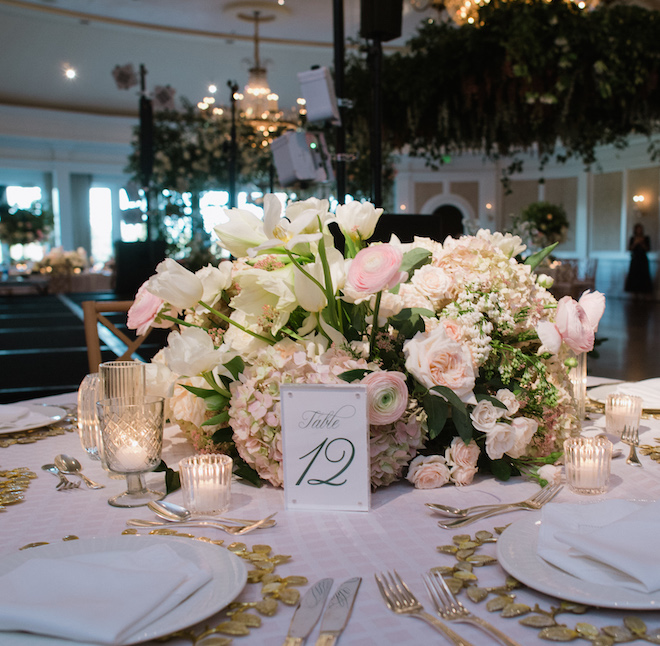 A floral centerpiece with a "Table 12" sign. The table has white plates with gold placemats and candlelight. 