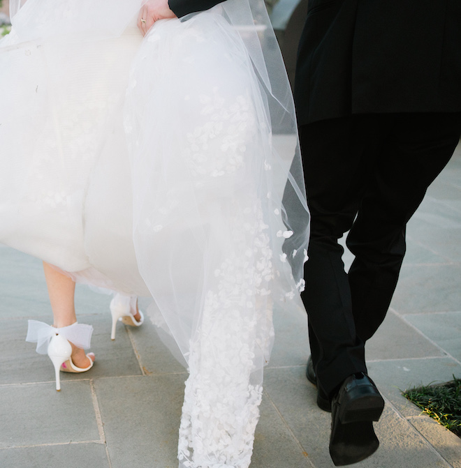 The bride and groom's feet walking down the sidewalk. The groom is holding the back of the bride's gown showing her white bow heels. 
