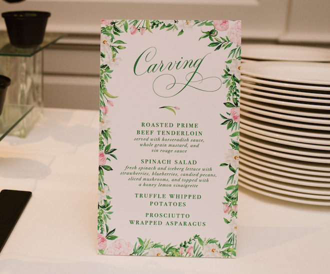 A white sign with pink and green florals. The sign has green writing reading the "Carving" station with four dishes below it. 