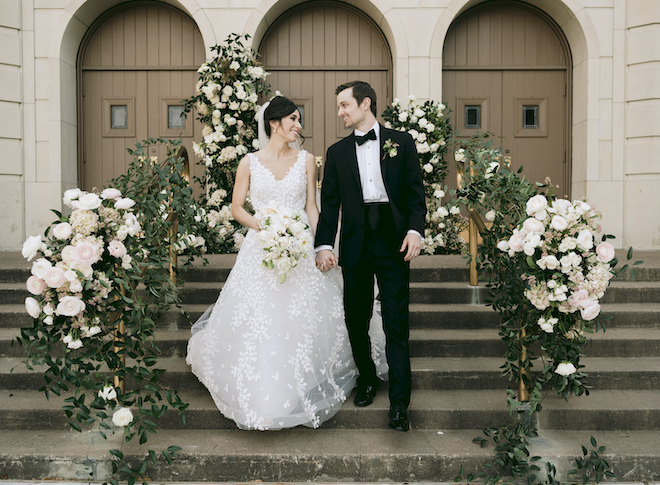 The bride and groom holding hands outside of the church with greenery and white florals decorating the stairs. 