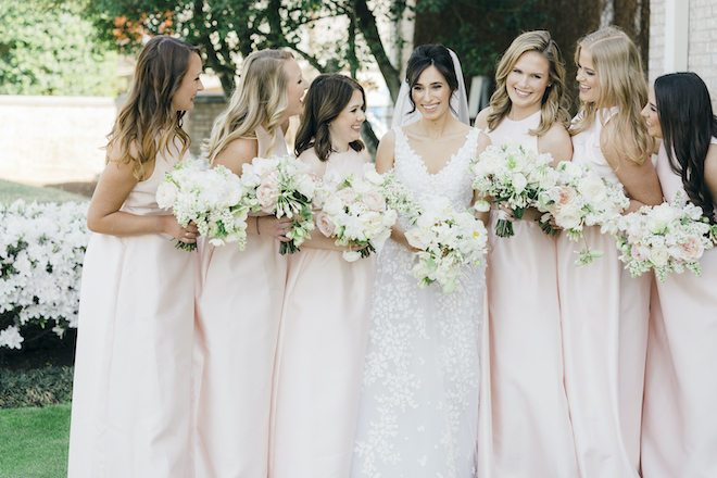 The bride smiling with her bridesmaids wearing light pink gowns and holding bouquets with white and pink florals. 